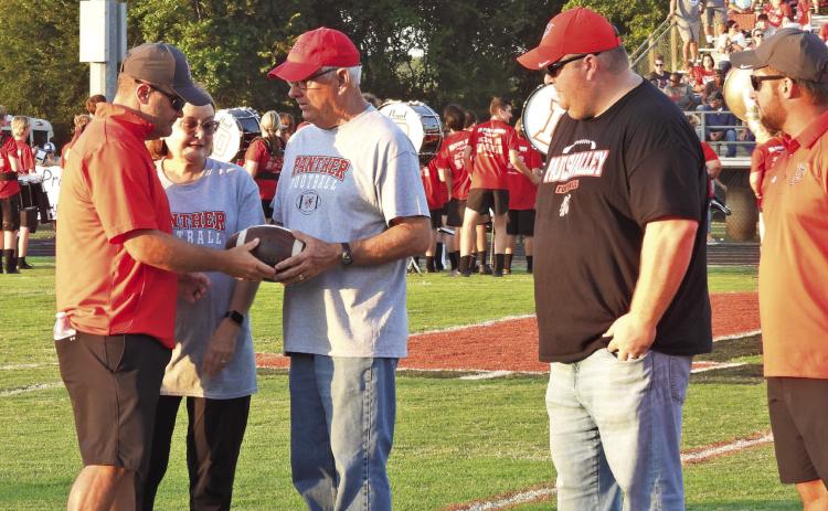 Pauls Valley head coach Dusty Raper (far left) hands a football signed by both Pauls Valley and Davis coaching staff to his father, retired longtime Davis Wolves Coach Jerry Don Raper, before Friday’s game to commemorate his influence on both programs. News Star photo by Chris Mackey